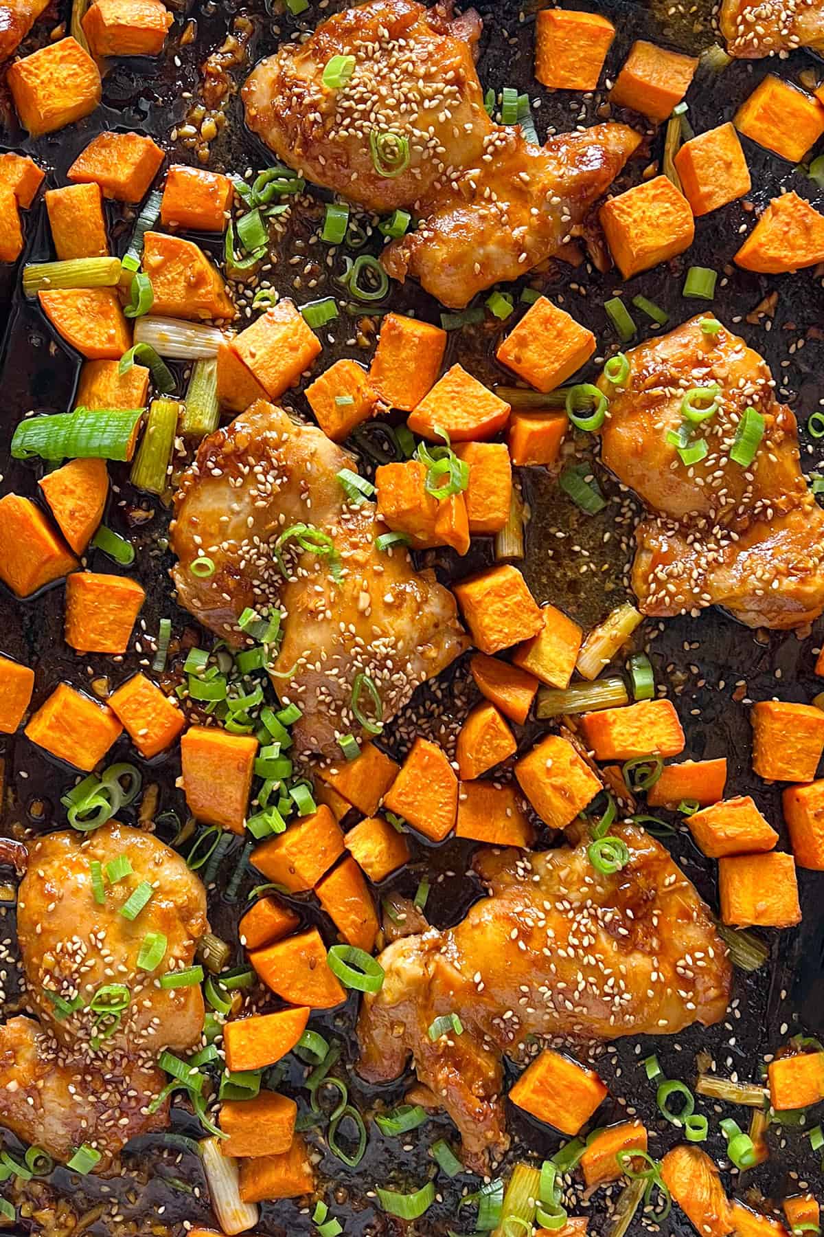 Sheet pan filled with roasted boneless chicken thighs and sweet potatoes cubes, topped with toasted sesame seeds and sliced scallions.