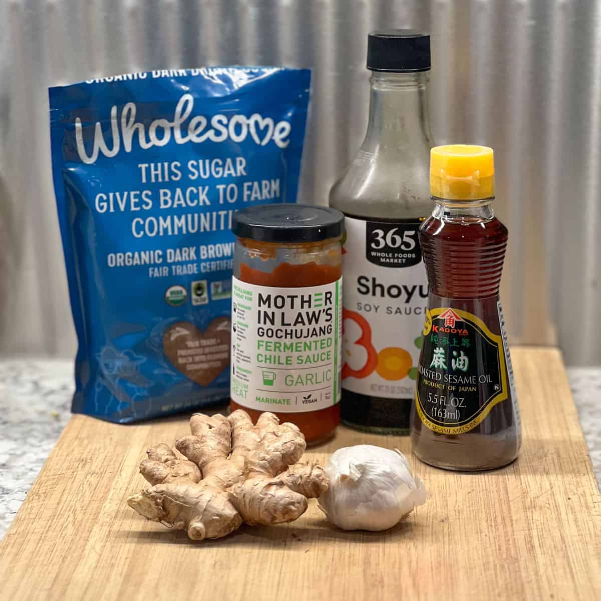 Ingredients on a wooden cutting board, bag of brown sugar, bottles of soy sauce, gochujang, and sesame oil, hunk of ginger root, whole garlic bulb.