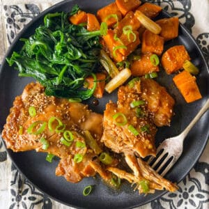 Two baked chicken thighs topped with scallions and sesame seeds, with roasted sweet potato cubes and sauteed spinach on a plate.