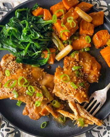 Two baked chicken thighs topped with scallions and sesame seeds, with roasted sweet potato cubes and sauteed spinach on a plate.
