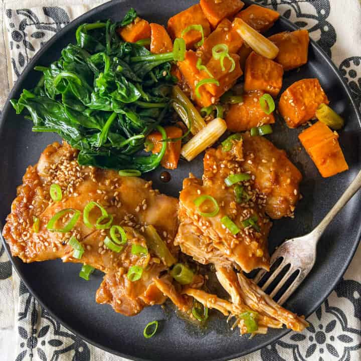 Two baked chicken thighs topped with scallions and sesame seeds, with roasted sweet potato cubes and sauteed spinach on a plate.