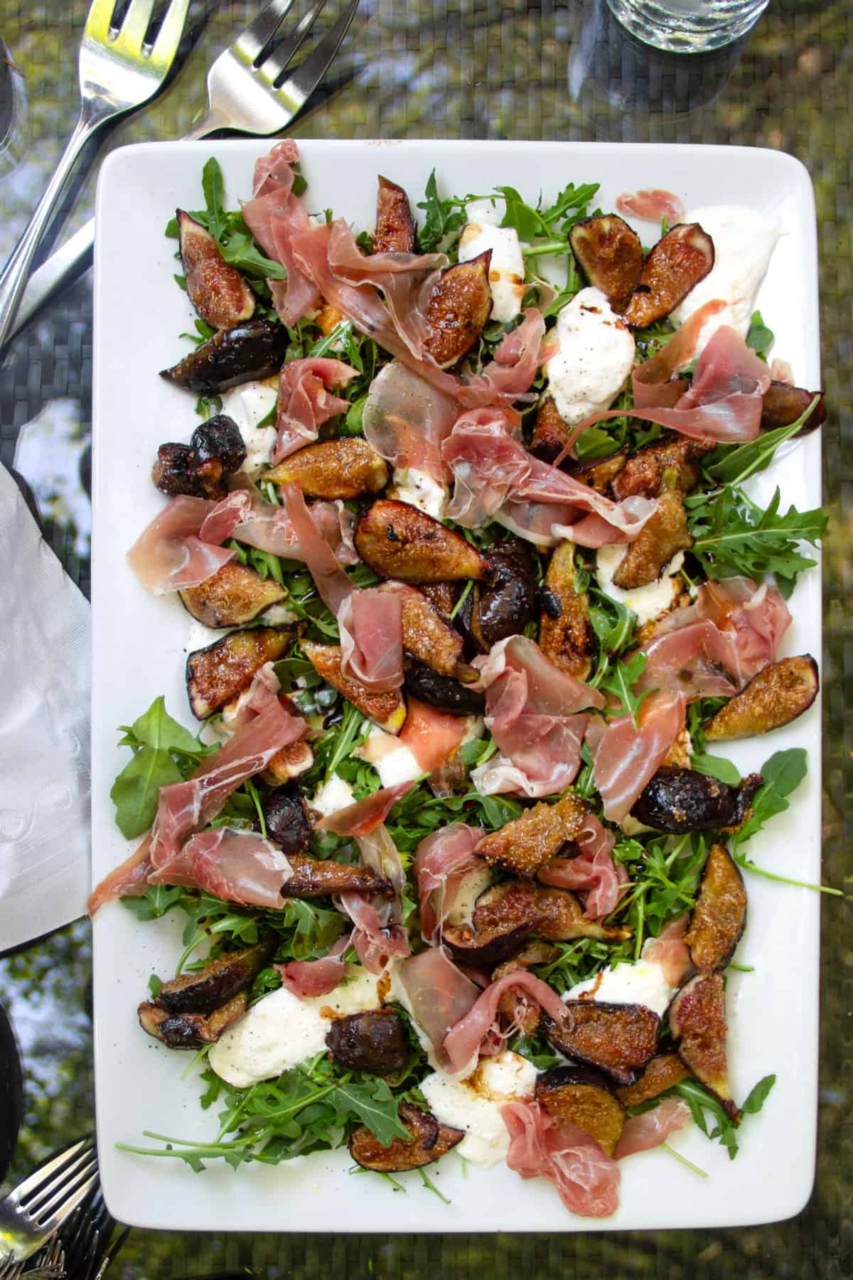 grilled figs, prosciutto and burrata arranged on a bed of arugula on a rectangular platter.