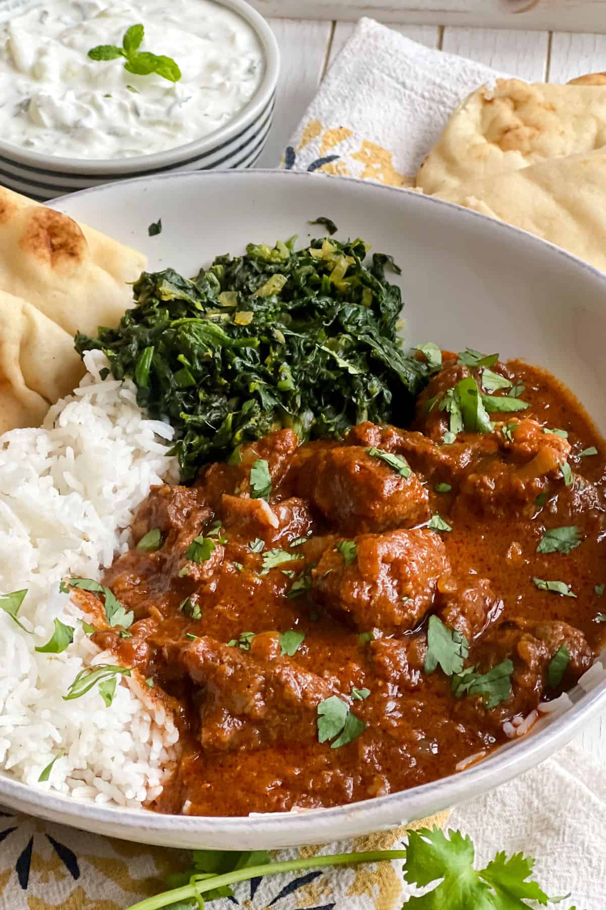 shallow bowl filled with lamb Rogan Josh with a side of sautéd spinach, white rice, and naan.
