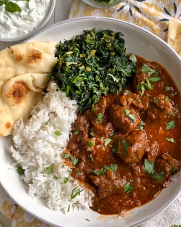 shallow bowl of lamb Rogan Josh with a side of spinach and rice and naan in the bowl.