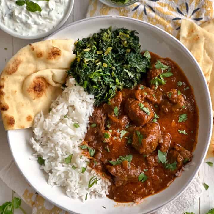 shallow bowl of lamb Rogan Josh with a side of spinach and rice and naan in the bowl.