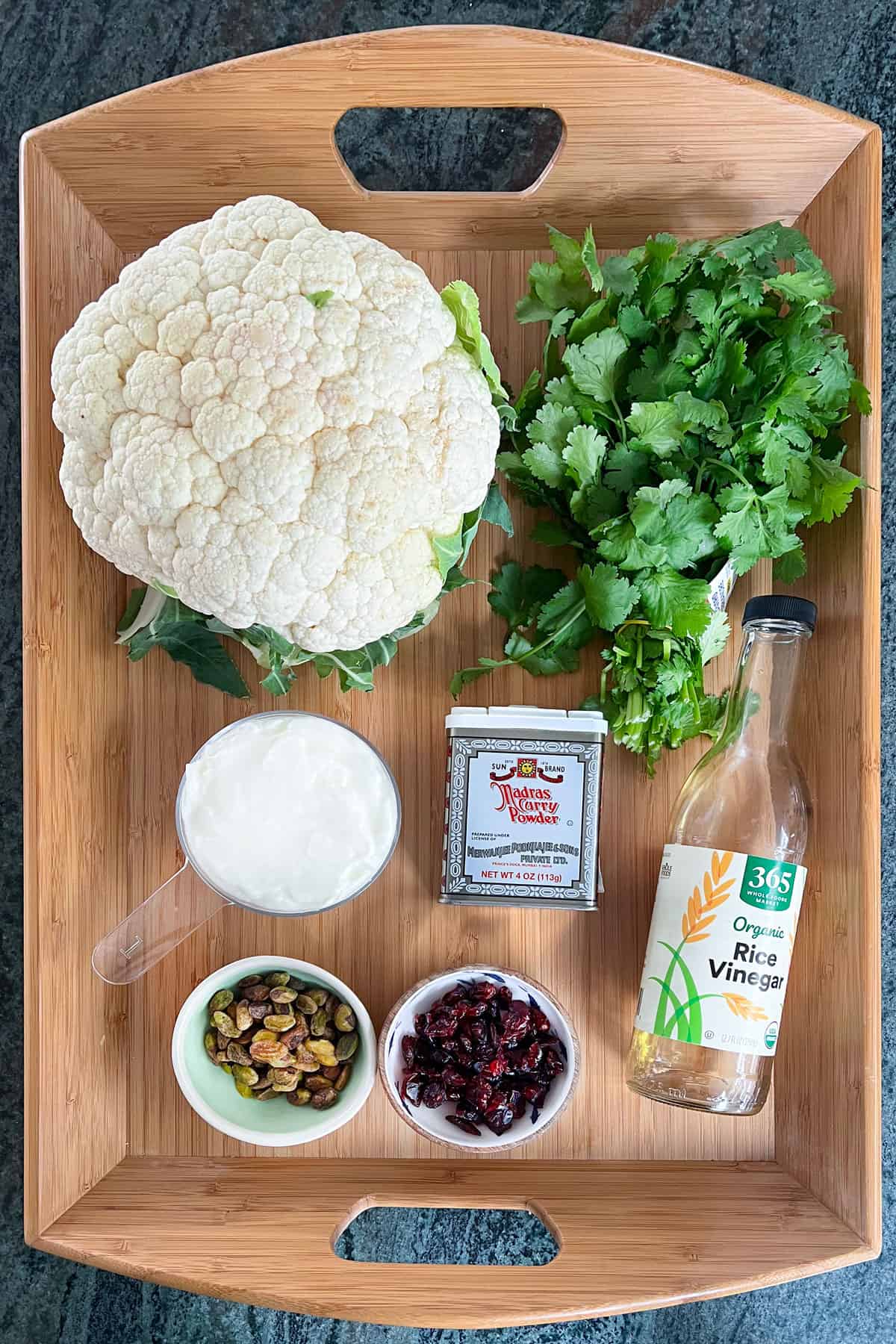 Ingredients for curried roasted cauliflower with yogurt and dried cranberries, set on a wooden tray: a head of cauliflower, a bunch of cilantro, a container of Madras curry powder, bowl of plain yogurt, bottle of rice vinegar, small bowls of pistachios and dried cranberries.