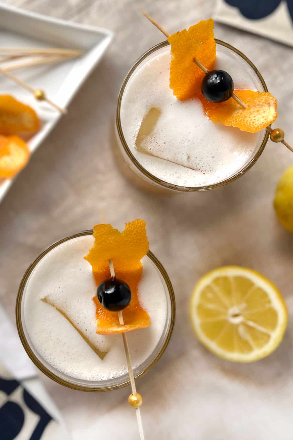 two whiskey sours on a table along with garnishes, lemons and toothpicks.