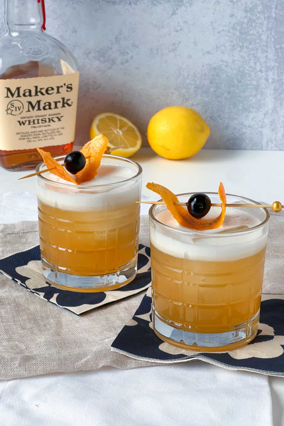 two whiskey sours on cocktail napkins with lemons and makers mark whiskey in the background.