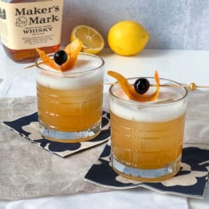 two whiskey sours on cocktail napkins with lemons and makers mark whiskey in the background.