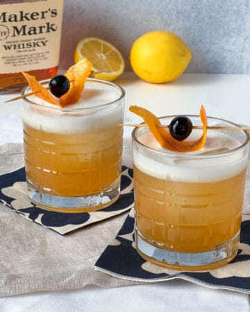 two whiskey sours on cocktail napkins with lemons and makers mark whiskey in the background.