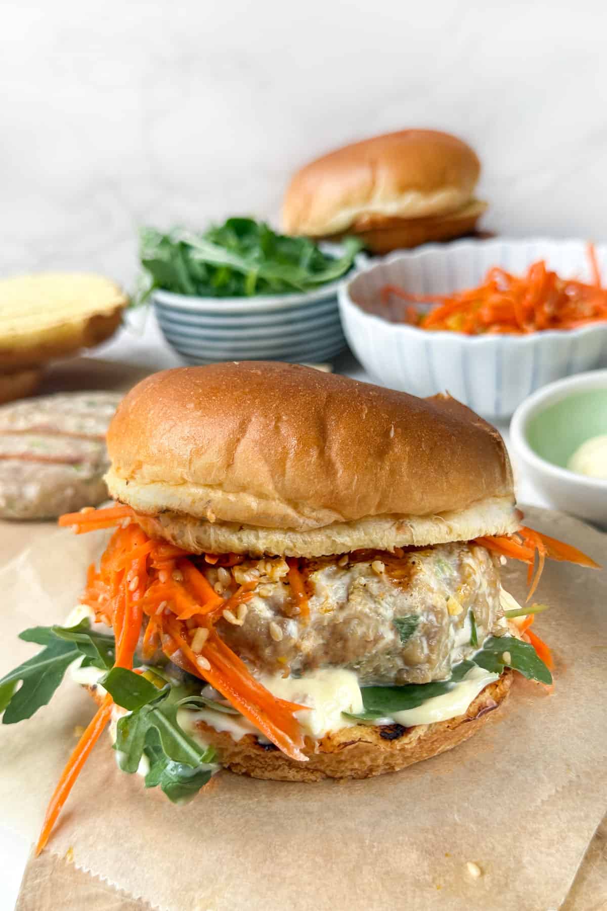 Ahi tuna burger with carrot slaw, arugula and kewpie mayo on a bun, with bowls of slaw, mayo and arugula in the background.