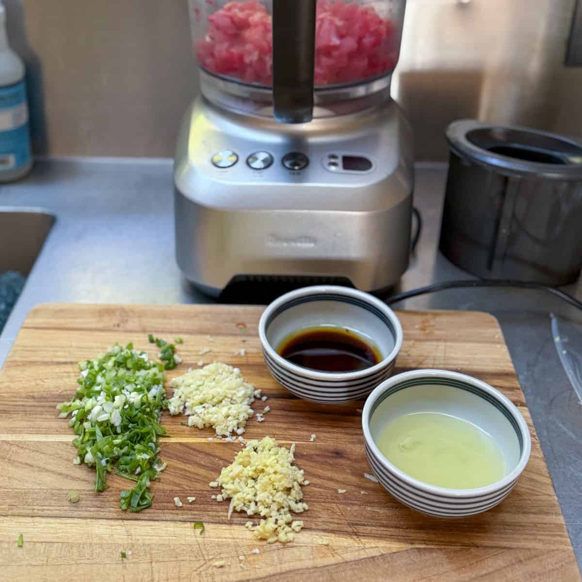 Small bowls of soy sauce and sesame oil, and chopped aromatics on a wooden cutting board, a food process in the background contained chopped ahi tuna.