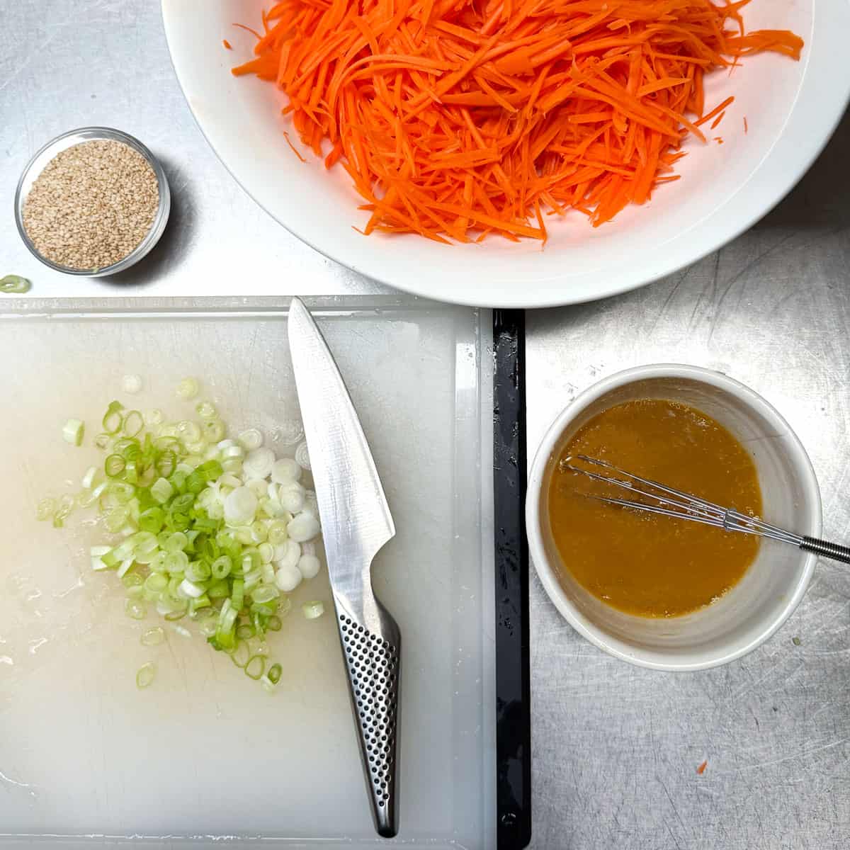 sliced scallions on a cutting board, a small bowl of sesame seeds, a medium bowl of ginger dressing and a bowl of shredded carrots.