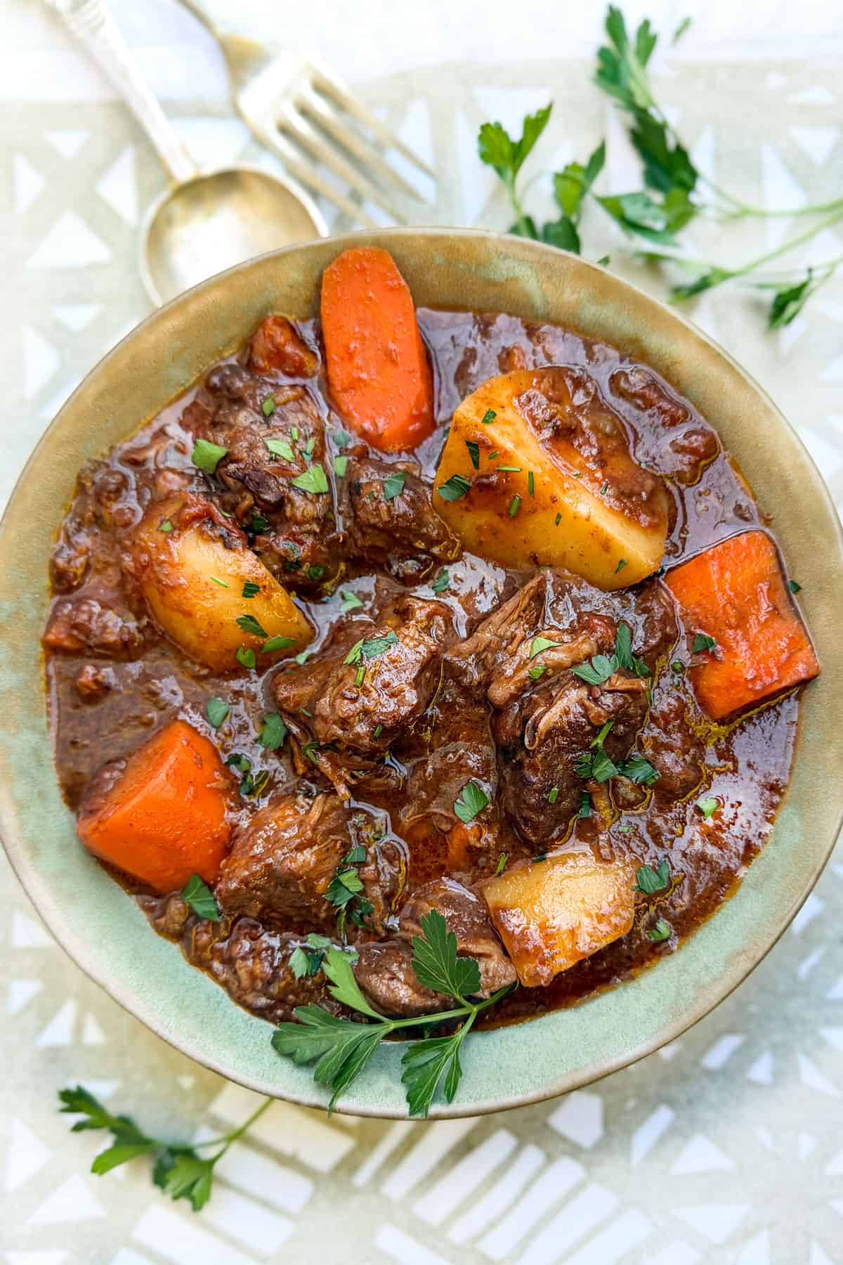 Beef stew with chunks of beef, carrots and potatoes in a bowl with a spoon and fork on the side and sprigs of parsley on the side.