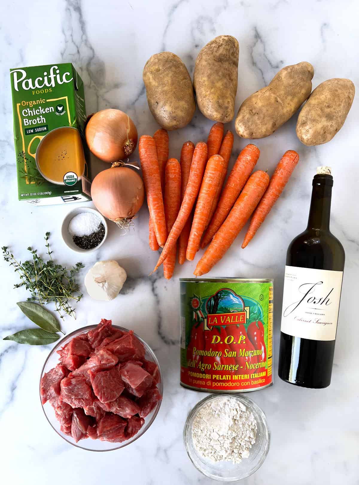 Ingredients for beef stew on a white marble countertop: container of chicken broth, 4 baking potatoes, two onions, bunch of carrots, sprigs of thyme, head of garlic, two bay leaves, can of plum tomatoes, bottle of red wine, bowl of cubed meat, bowl of white flour.