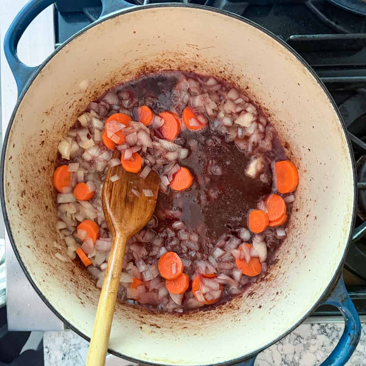 carrots, onions and red wine in dutch oven and being stirred with a wooden spoon.